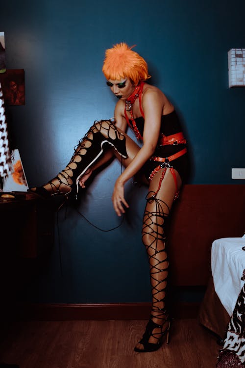 Free Side view full body bizarre female with dark makeup and orange hair wearing black bodysuit and leather BDSM harness putting leg on vanity table and lacing thigh high shoe Stock Photo
