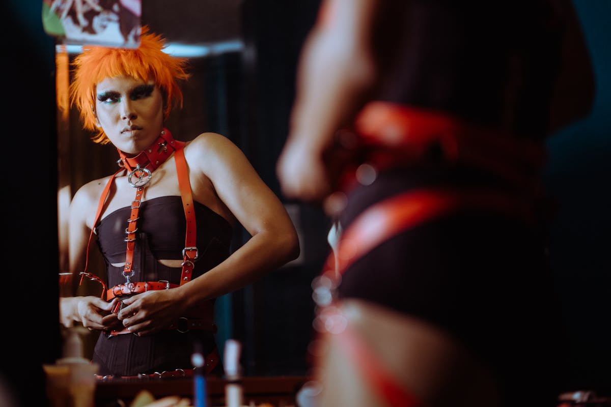 Crop informal female with funky makeup and orange hair wearing black bodysuit and putting on leather BDSM harness while looking in mirror
