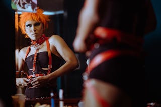 Crop informal female with funky makeup and orange hair wearing black bodysuit and putting on leather BDSM harness while looking in mirror