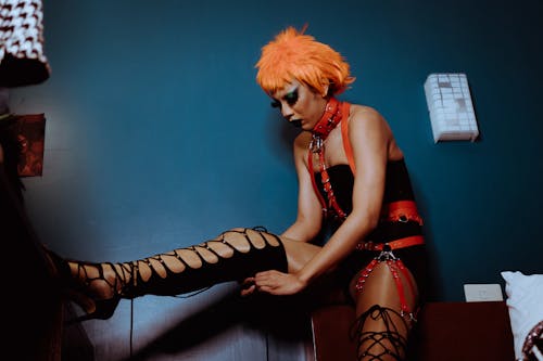 Free Extravagant female with dark makeup and orange wig wearing black bodysuit and leather harness raising leg on vanity table and zipping thigh high shoe in dark bedroom Stock Photo