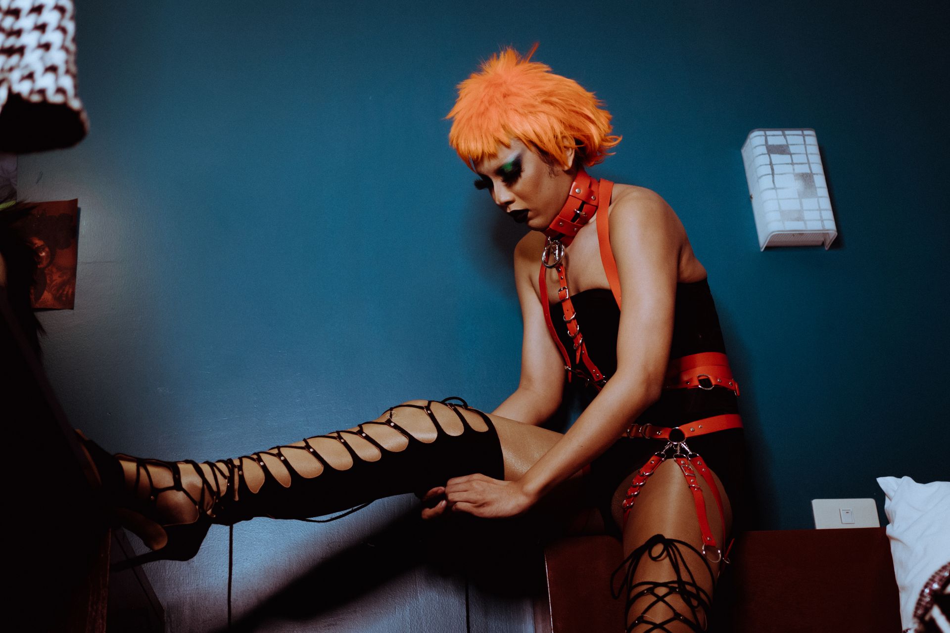 Extravagant female with dark makeup and orange wig wearing black bodysuit and leather harness raising leg on vanity table and zipping thigh high shoe in dark bedroom