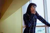 Thoughtful female with unusual dark makeup wearing warm dress smoking cigarette and looking out window pensively in hallway in daylight