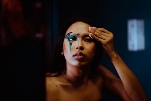 Unemotional ethnic female with bare shoulders reflecting in mirror while removing dark body art makeup from face by using cleansing wipe in dark studio