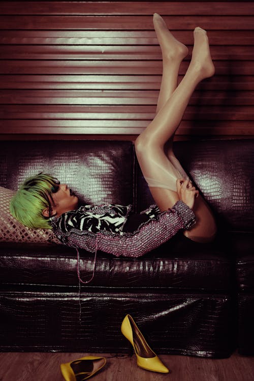 Free Side view full length transsexual informal female in green wig and short dress lying on leather couch with legs raised and adjusting stockings Stock Photo