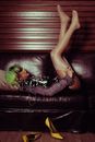 Side view full length transsexual informal female in green wig and short dress lying on leather couch with legs raised and adjusting stockings