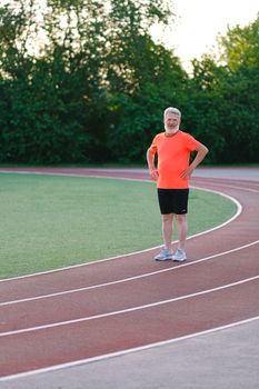 Sporty bearded man in orange t shirt and black shorts holding hands on waist looking at camera while standing on running track