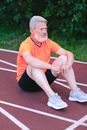 Side view of senior male athlete in bright t shirt and black shorts looking away having rest while sitting on stadium track