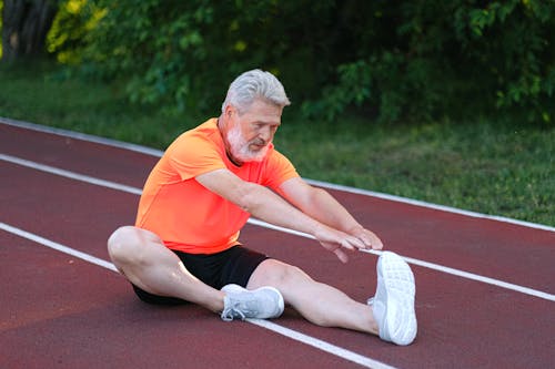 Full body of focused senior male sitting and stretching on racetrack in stadium near bushes with green leaves