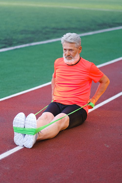 Free Full body of elderly male stretching and warming up with elastic rubber on racetrack in stadium Stock Photo