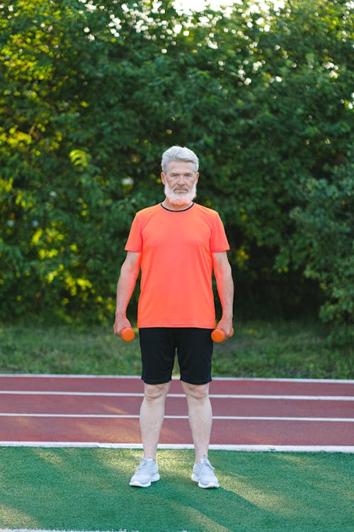 Full body of focused elderly man with beard in sportswear standing with dumbbells on stadium and looking at camera
