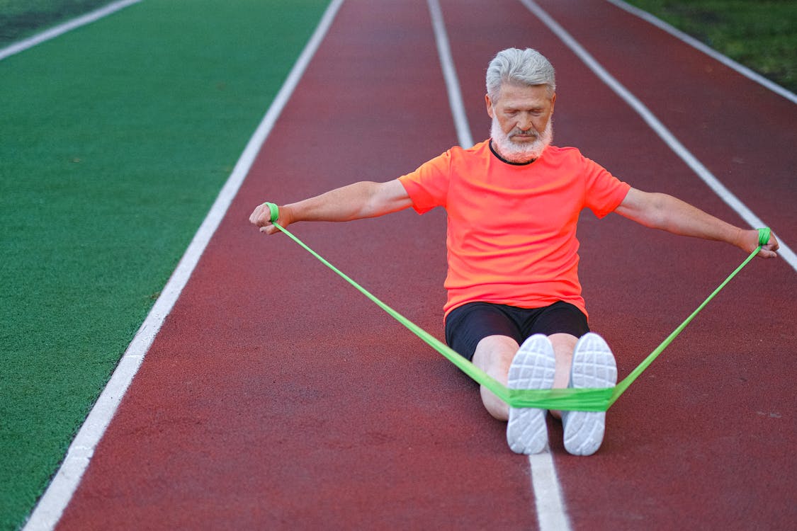 Concentrate elderly sportsman sitting on sport running track on stadium and doing dynamic workout pulling rubber elastic in daytime