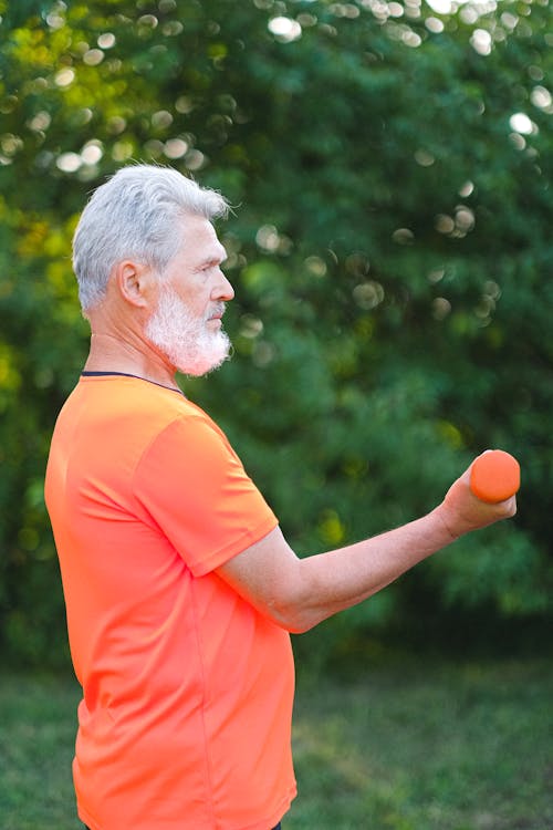 Side view of active senior man in bright sportswear training biceps with dumbbells in park in daytime