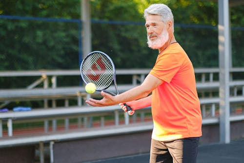 Side view of cheerful senior sportsman standing with ball and racket and preparing to serve ball on court in daytime