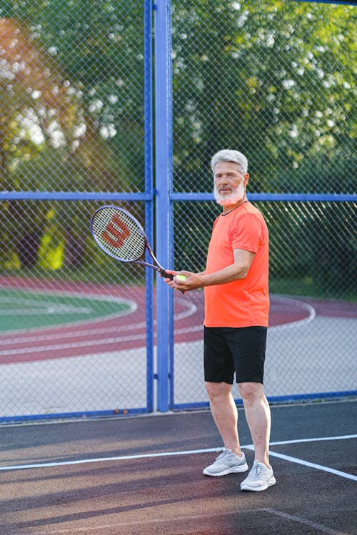 Free Concentrated aged tennis player in sportswear standing with racket and ball during tennis match on sports ground in sunny day Stock Photo