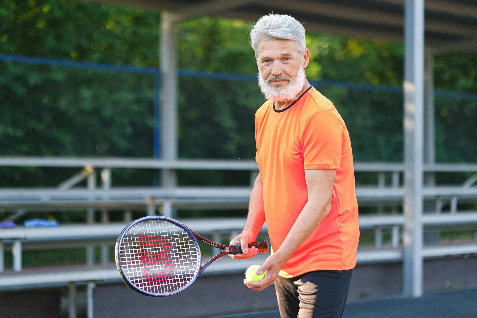 Positive senior sportsman in bright t shirt preparing to hit ball during tennis training on court in sunny day