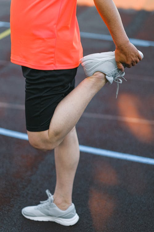 Side view of crop anonymous athlete in sportswear standing on red running track and stretching legs before training on stadium in daytime