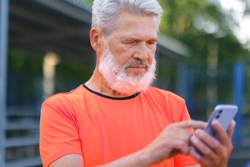 Crop elderly bearded male with gray hair browsing modern mobile phone while spending time in park