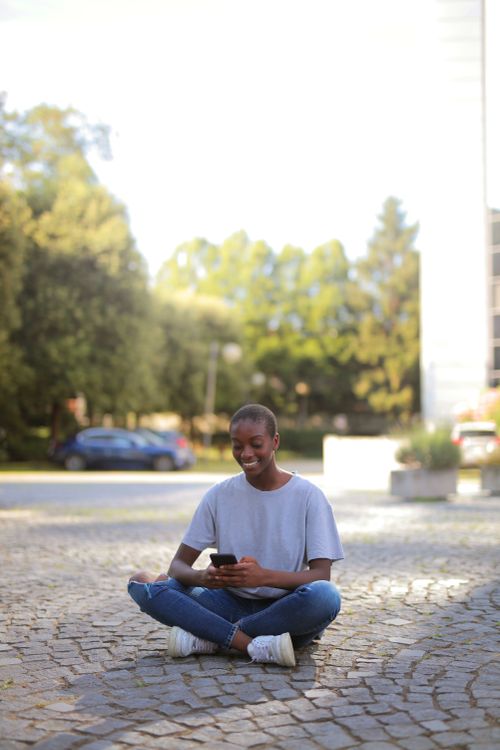 Free Happy teenager sitting on a cobblestone street using a smartphone, enjoying a sunny day. Stock Photo