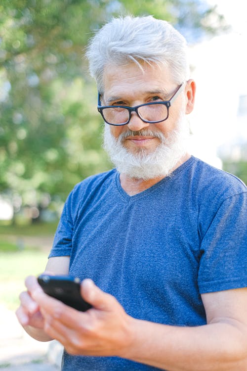 Elderly bearded male with gray hair in glasses and casual clothes standing in park and browsing mobile phone while looking at camera