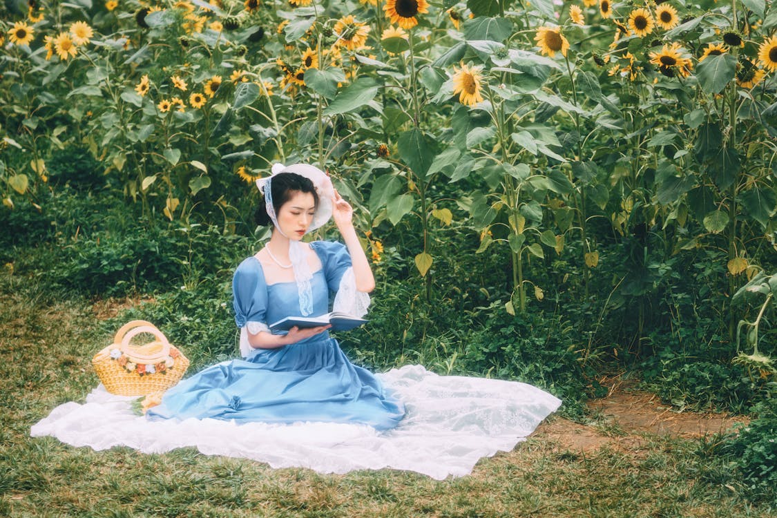 Girl in Blue Dress Sitting on White Textile Surrounded by Green Plants
