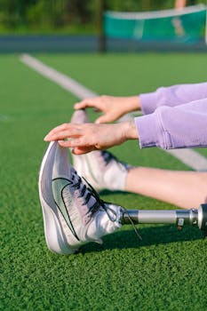 Crop unrecognizable female athlete with leg prosthesis doing stretching exercises while training on green court