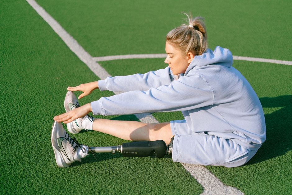 Full body side view of handicapped female with leg prosthesis sitting on grassy sports ground while stretching body before training
