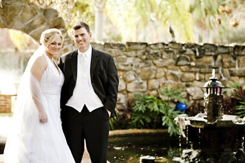 Man and Woman in Their Wedding Outfit With Brown Wall in the Background Near Fountain and Pond during Daytime
