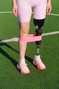 Unrecognizable female athlete with leg prosthesis standing on grassy sports ground with rubber expander while practicing stretching exercises on stadium