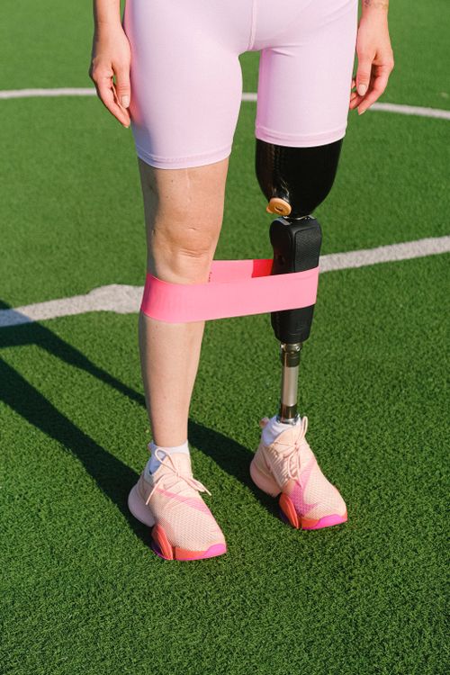 Unrecognizable female athlete with leg prosthesis standing on grassy sports ground with rubber expander while practicing stretching exercises on stadium