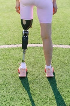 Back view of a female athlete with a prosthetic leg standing on a sunny sports field, showcasing strength and determination.