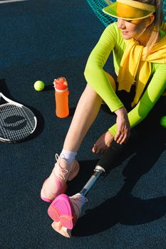 Young woman with prosthetic leg sitting on a tennis court, embracing active lifestyle in bright sportswear.