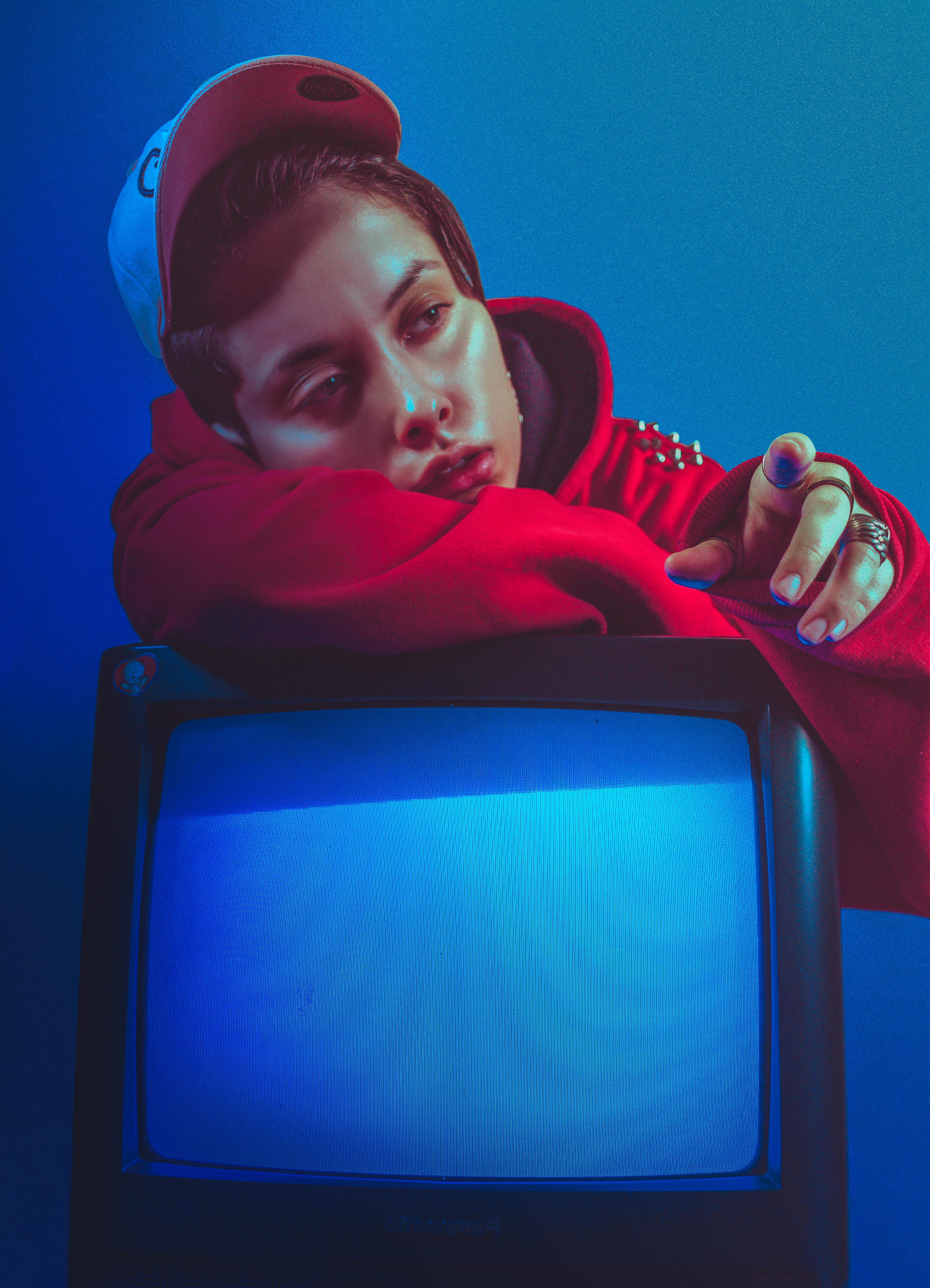 Stylish portrait of a person in a red hoodie and cap resting on a vintage TV.