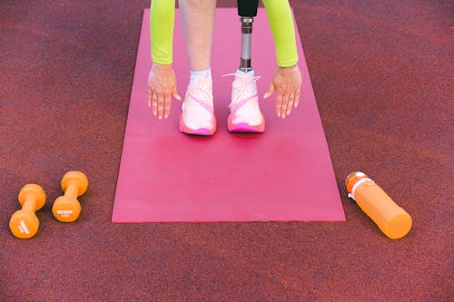Unrecognizable female athlete with leg prosthesis bending forward and stretching while trying ti touch ground during training on sports ground with dumbbells
