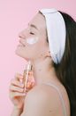 Side view of cheerful female with headband and face cream standing with eyes closed on pink background with face oil