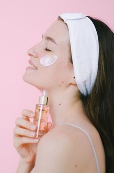 Side view of cheerful female with headband and face cream standing with eyes closed on pink background with face oil