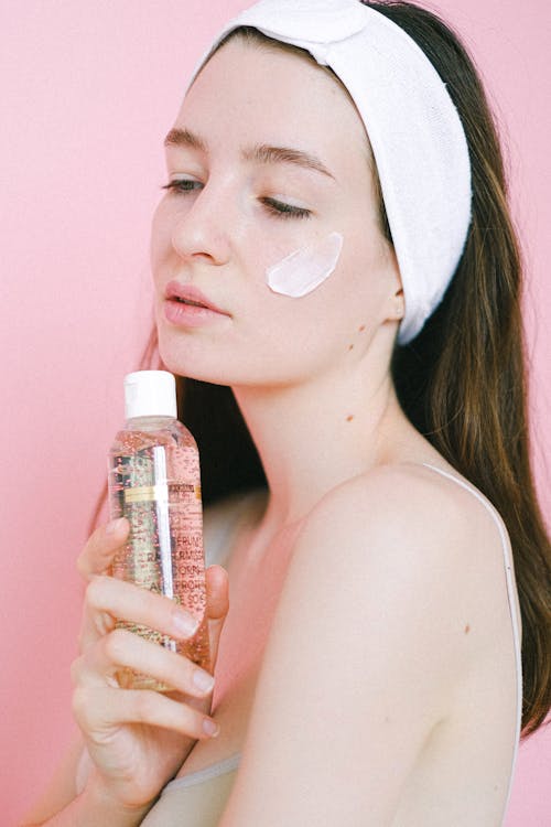 Calm female wearing headband with face cream on cheek standing on pink background with bottle of cosmetic tonic during skincare