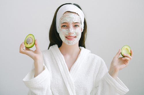 Free Cheerful female with facial mask wearing bathrobe and headband looking at camera while standing on white background with avocado in hands Stock Photo