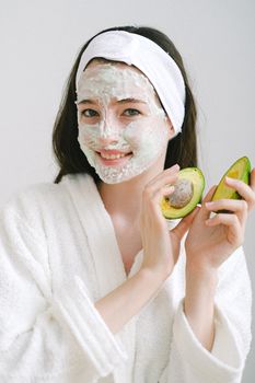 一位乐观的女性脸上涂着保湿霜，身穿浴衣，手拿牛油果站在白色背景前看着镜头