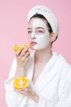 Woman in a bathrobe, applying face mask, holding an orange on a pink background.