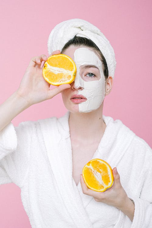 Calm female with towel on head and facial mask on face looking at camera while standing on pink background and covering eye with half of orange