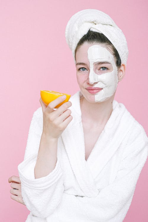 Cheerful female with towel on head and facial mask on face looking at camera while standing on pink background with arm bent in elbow and orange in hand