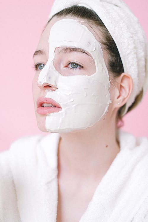 Serene female in bath robe and towel on head standing with applied facial mask on half face in light spa salon and looking away