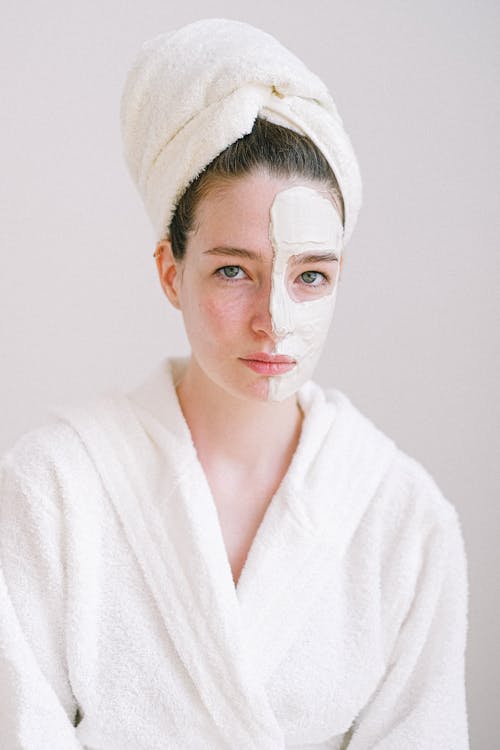 Young attractive female in white bathrobe and towel on head standing in spa salon with rejuvenating facial mask on half face and looking at camera