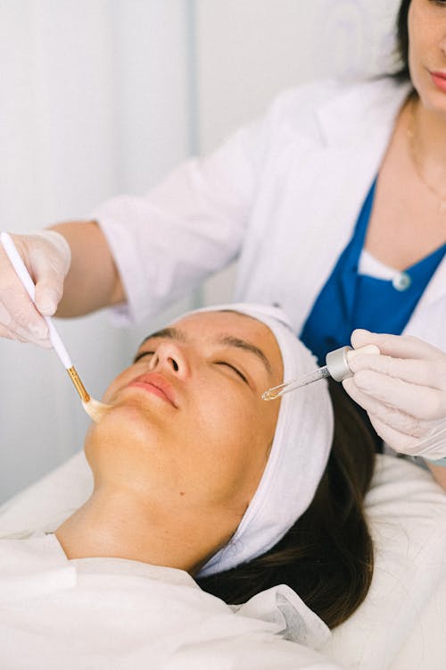 Free Young female cosmetologist in medical robe applying rejuvenating essential oil on female customer face skin lying with eyes closed in light modern spa salon Stock Photo