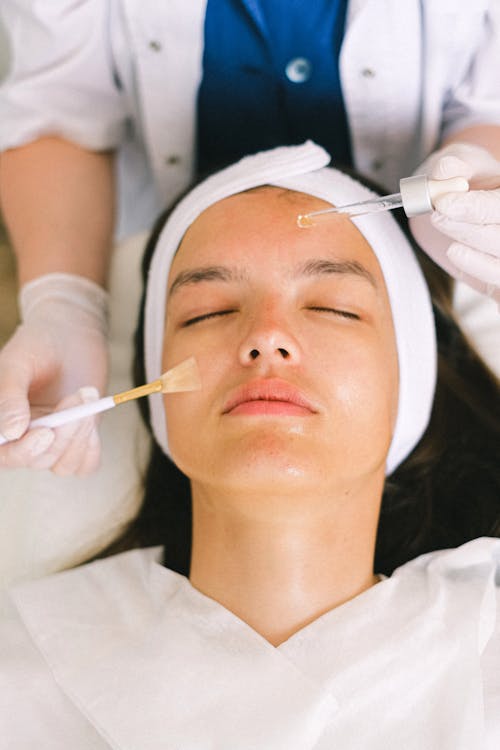 Free Top view skilled female cosmetologist in uniform and gloves applying essential oil on young woman face skin while working in contemporary spa center Stock Photo