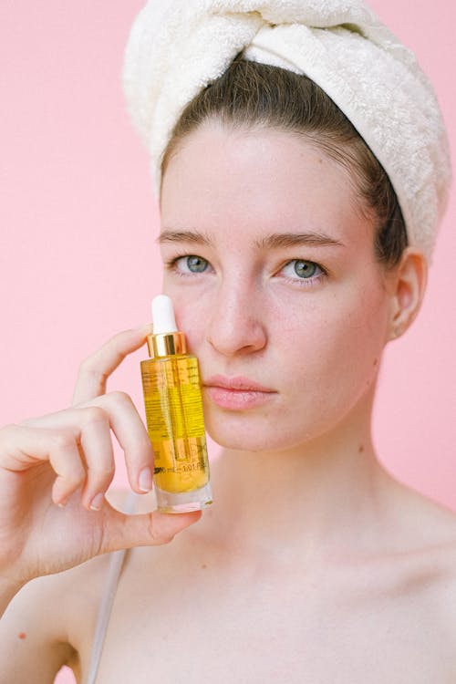 Crop young female without makeup and with hair wrapped in towel showing cosmetic product holding bottle near pretty face and looking at camera