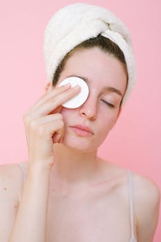 Young woman cleansing face with cotton pad on pink background