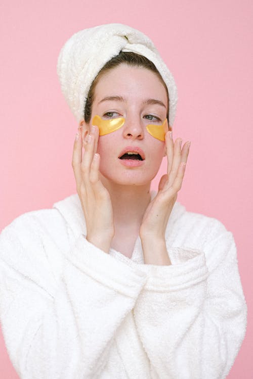 Beautiful female in white bathrobe and towel on head applying revitalizing eye patches and touching face while looking away against pink background
