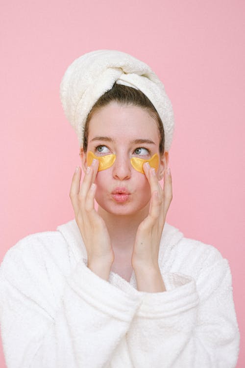 Attractive young female wearing white bathrobe and towel on head applying eye patches on face and pouting lips while looking away against pink background