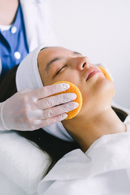 Female beautician in medical robe and gloves using yellow sponges to clean female customer face lying with eyes closed in spa